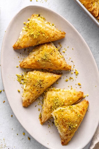 A few Shaabiyat on a long narrow platter garnished with crushed pistachio, baking dish with more still in square shape and a small dish of crushed pistachios nearby.
