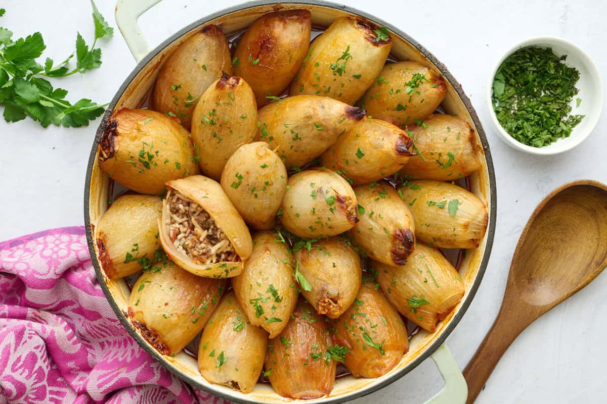 Stuffed onions filled with ground beef, rice and tomatoes in the skillet garnished with chopped parsley, one onion cut open and positioned upwards to see filling inside, small dish of parsley and serving spoon nearby.