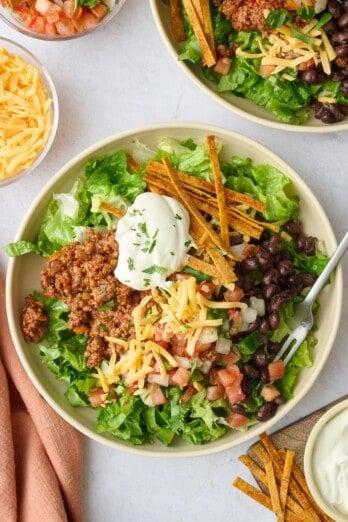 Easy taco salad recipe served in bowls with lettuce, ground beef taco meat, cheese, beans, pico de gallo, sour cream and tortilla strips.