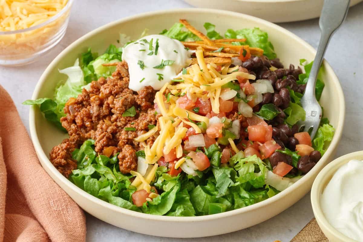 Best taco salad recipe served in bowls with lettuce, ground beef taco meat, cheese, beans, pico de gallo, sour cream and tortilla strips.
