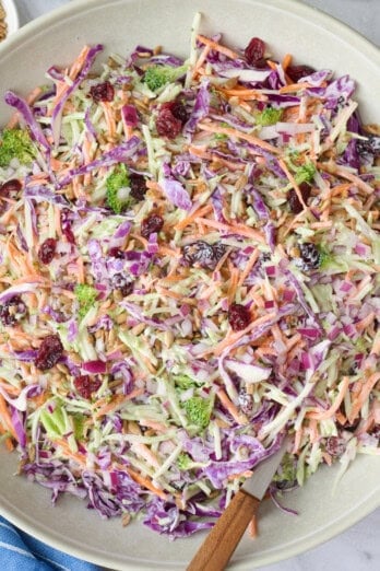 Broccoli slaw in a serving bowl with serving set dipped inside, small dishes of extra dried cranberries and sunflower seeds nearby.