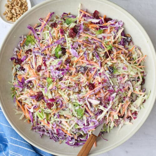 Broccoli slaw in a serving bowl with serving set dipped inside, small dishes of extra dried cranberries and sunflower seeds nearby.