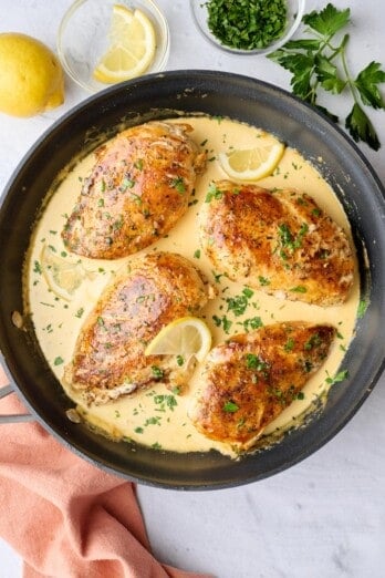 Creamy lemon chicken in skillet with sauce spooned lightly on top of each chicken, garnished with chopped parsley, small dish of lemons nearby.