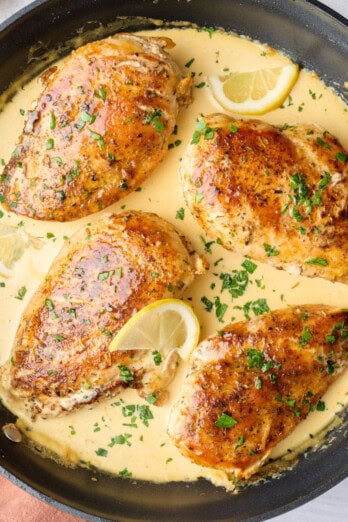 Creamy lemon chicken in skillet with sauce spooned lightly on top of each chicken, garnished with chopped parsley, small dish of lemons nearby.