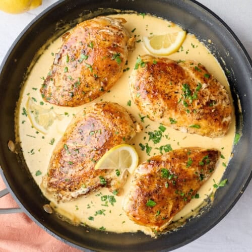 Creamy lemon chicken in skillet with sauce spooned lightly on top of each chicken, garnished with chopped parsley, small dish of lemons nearby.
