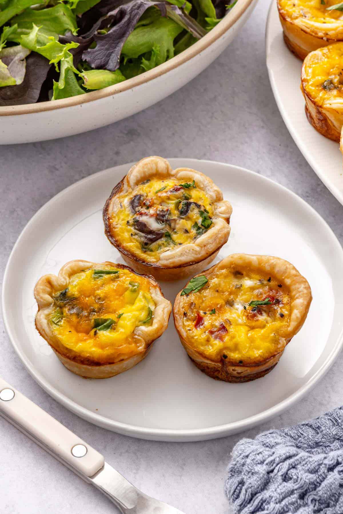 3 mini quiche on a small plate with spring mix salad, platter nearby.