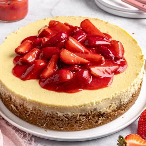 Whole strawberry cheesecake on a round platter with a small dish of extra strawberry sauce nearby.