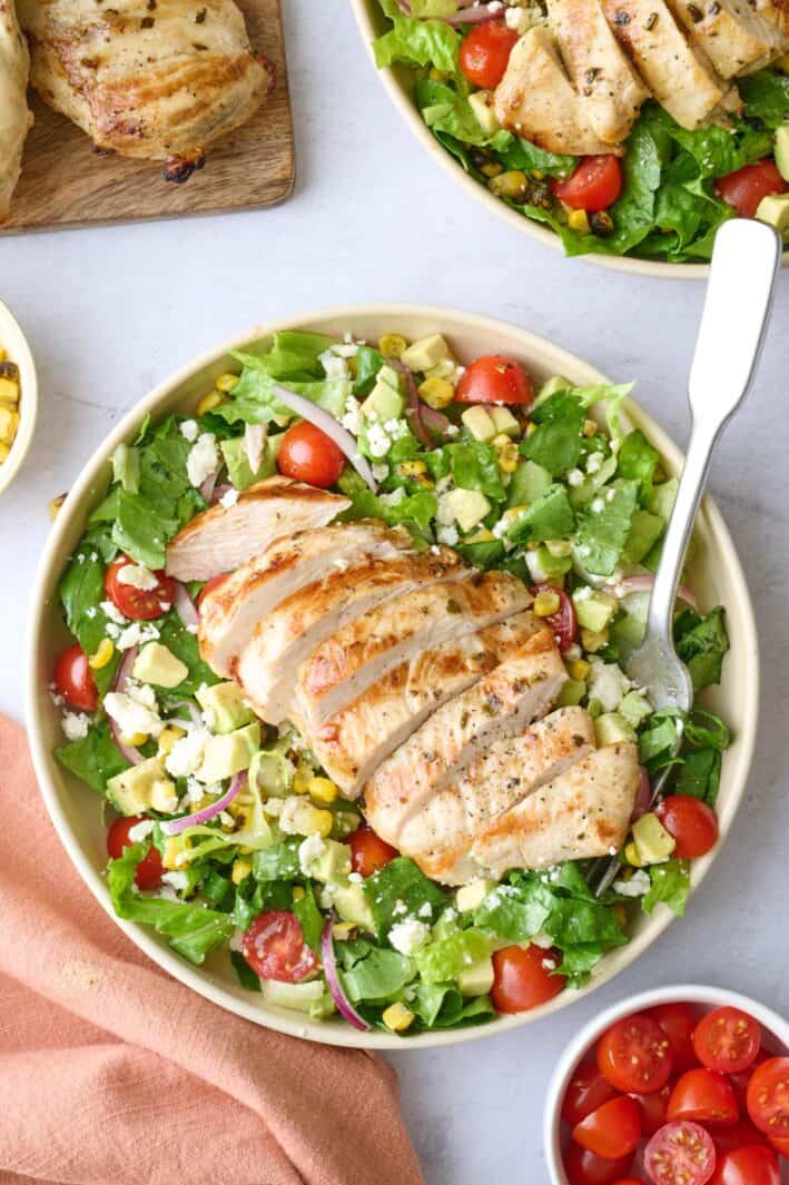 2 bowls of grilled chicken salad with extra fresh ingredients around.