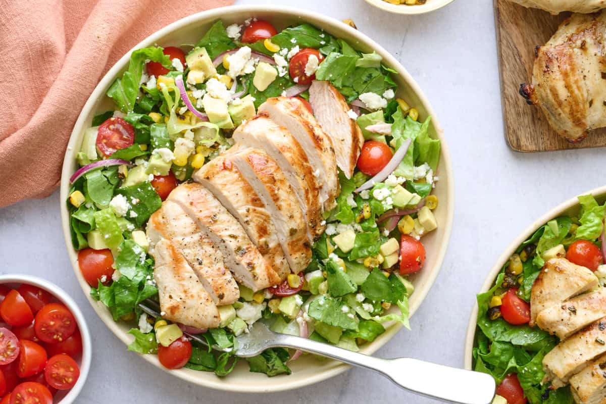 2 bowls of grilled chicken salad with extra fresh ingredients around.