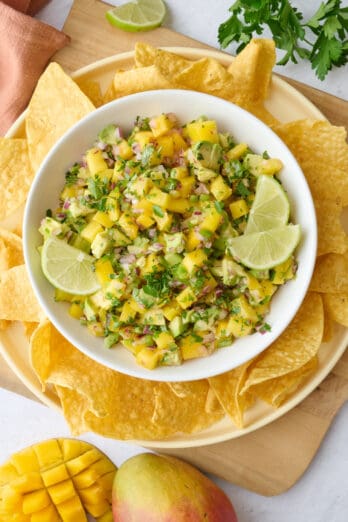 Mango salsa in a serving bowl garnished with lime wedges and extra cilantro, tortilla chips and a halved mango cut on the skin.