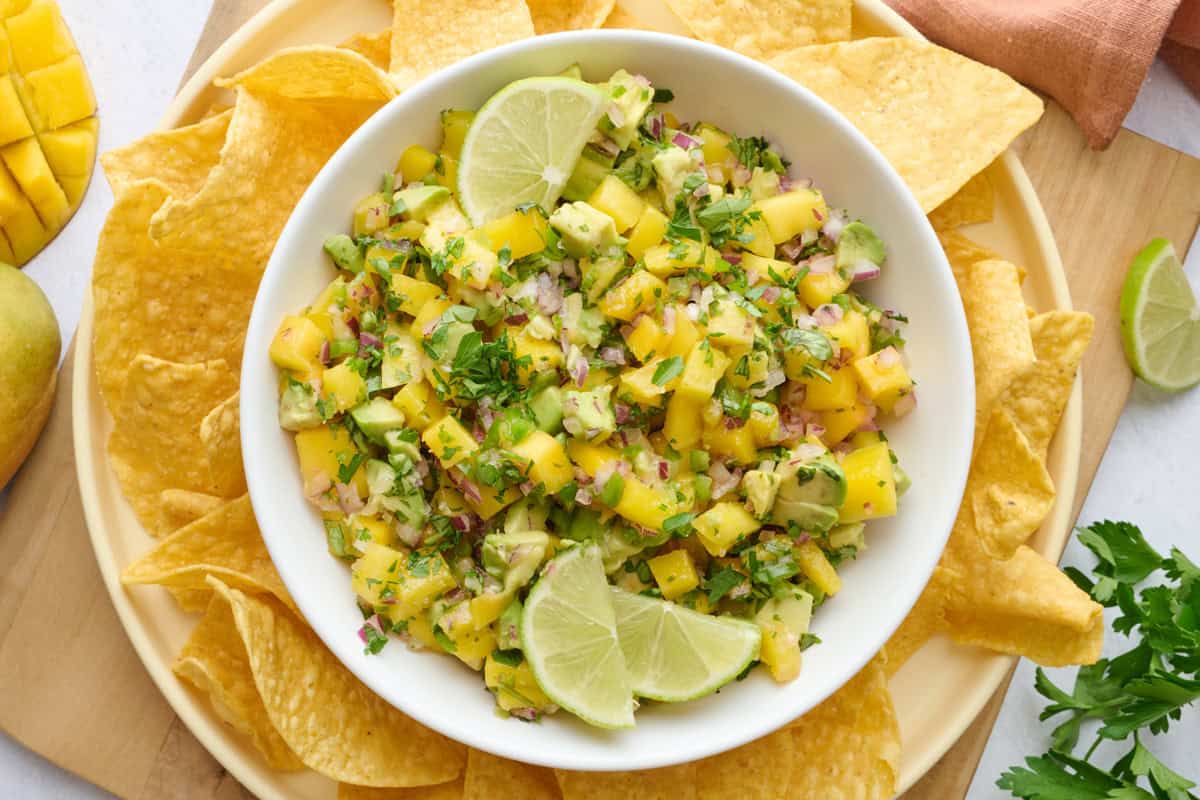 Mango salsa in a serving bowl garnished with lime wedges and extra cilantro, tortilla chips and a halved mango cut on the skin.