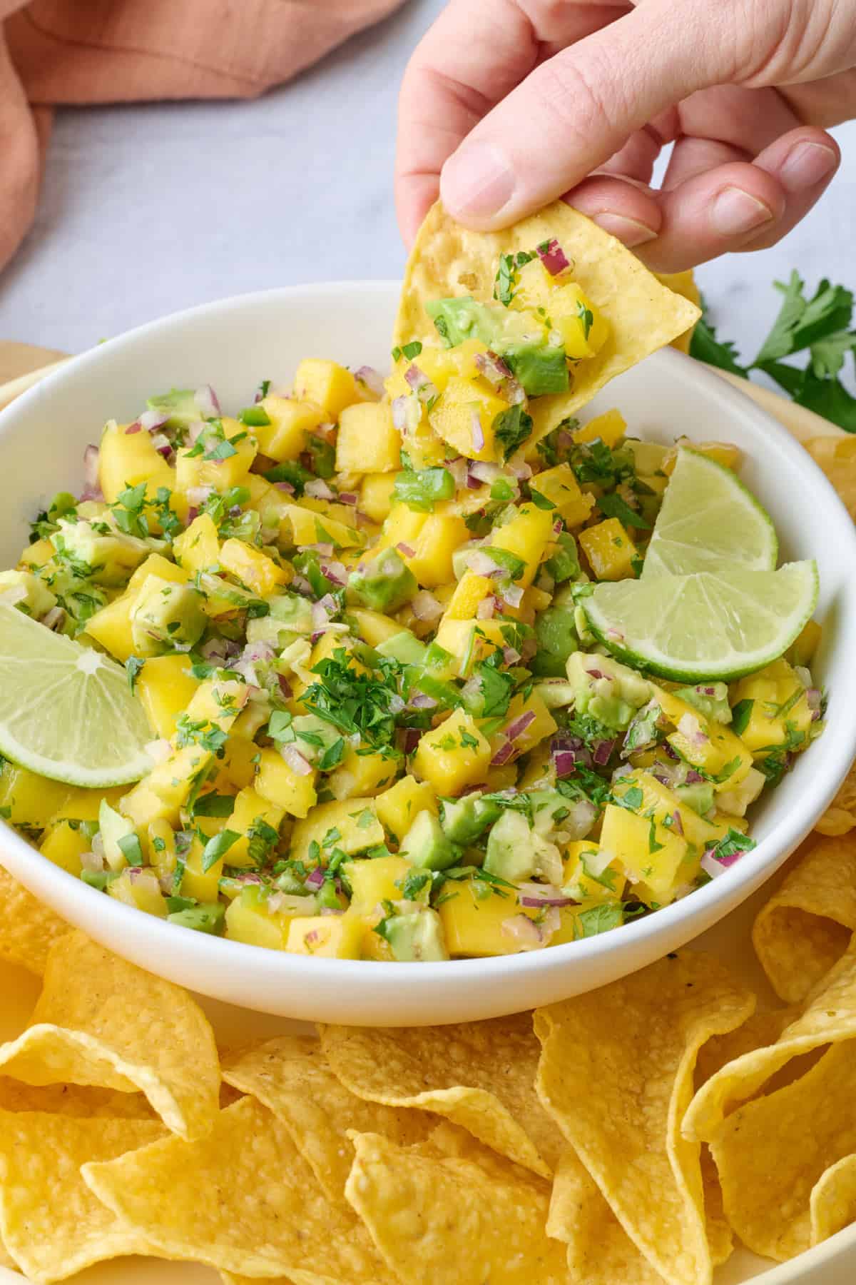Mango salsa in a serving bowl garnished with lime wedges and extra cilantro, with a tortilla chip dipped inside.
