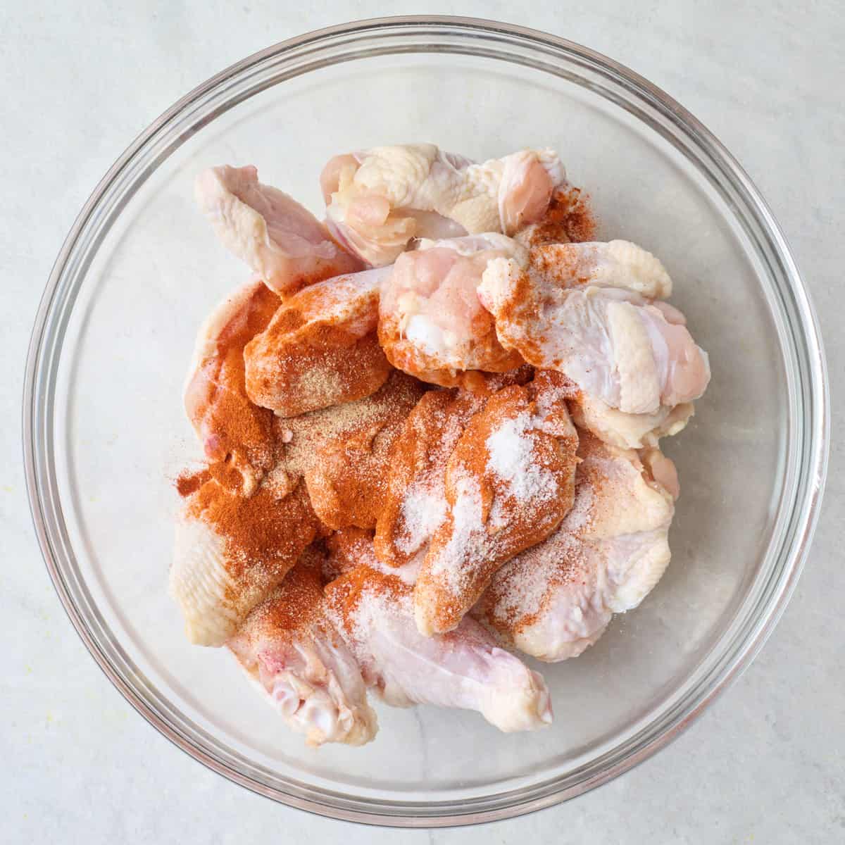 Chicken wings in a bowl with seasonings and oil added on top before tossing together.